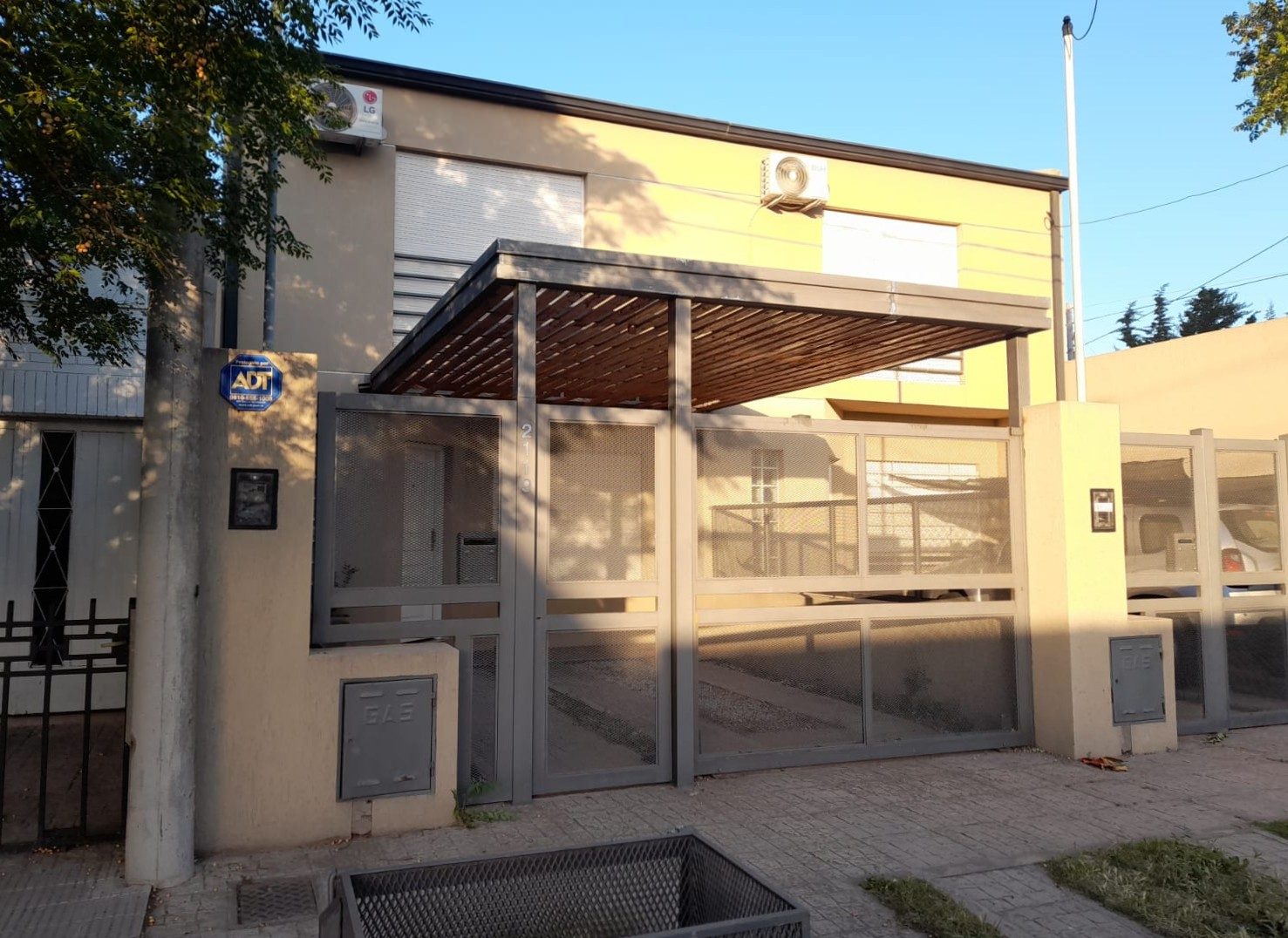 Pérgola de estructura metálica con listones de madera sobre un frente con portón de malla