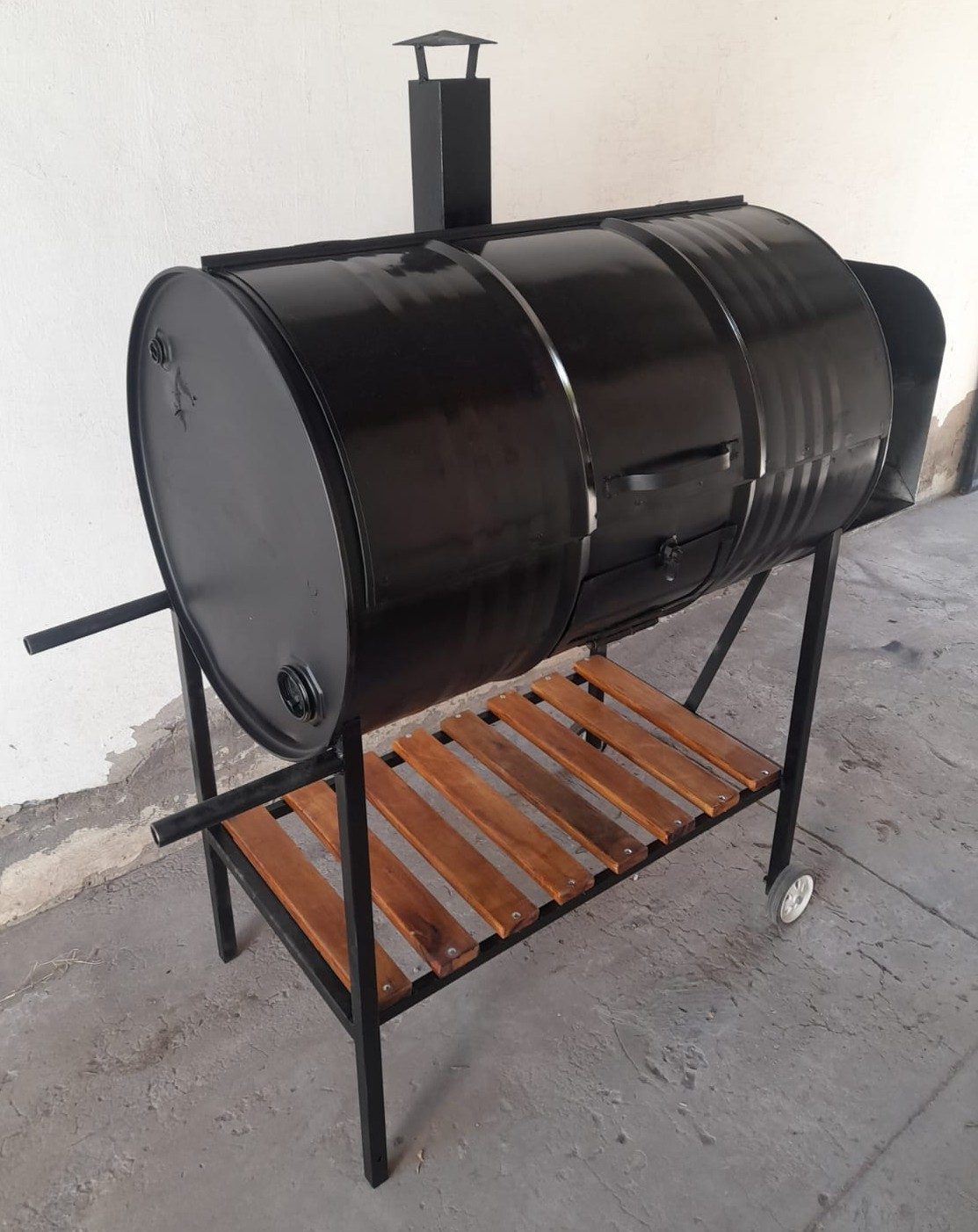 Ahumador cilíndrico de metal negro con chimenea, ruedas y estante inferior de madera
